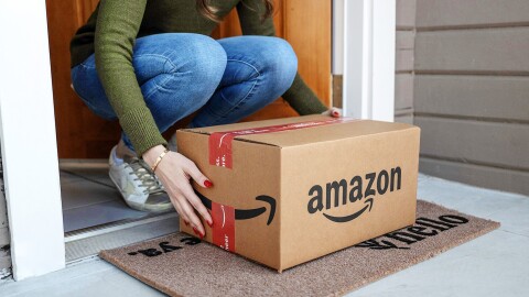 A person bends down to pick up an Amazon box delivered to a doorstep.
