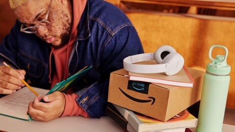 A man in a denim jacket opens a book to study; to his left on the table there's an unopened Amazon box, a mint green Takeya water bottle, and white Sony headphones.