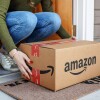 A person bends down to pick up an Amazon box delivered to a doorstep.